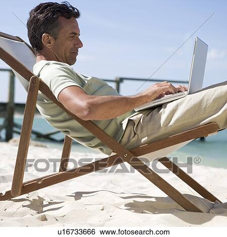 Stock Photograph - A mature man sitting in a deck chair using a laptop. Fotosearch