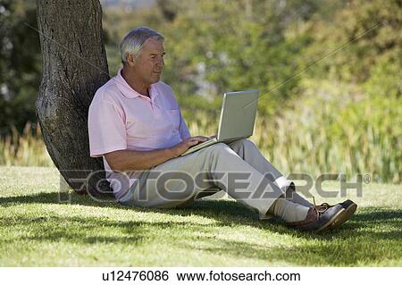 Stock Photograph - A senior man sitting under a tree using a laptop. Fotosearch