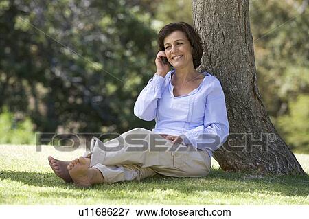 Stock Photo - A senior woman sitting under a tree using a mobile telephone. Fotosearch