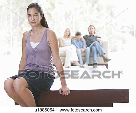 Stock Image - A young girl sitting on a coffee table with her family on the sofa. Fotosearch