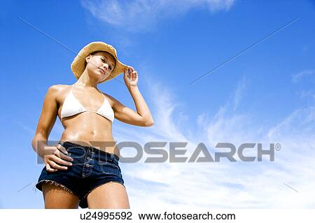 Stock Image - A young woman in bikini and shorts against blue sky. Fotosearch