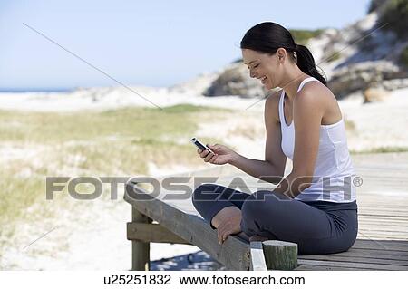 Stock Image - A young woman sitting on a beach using a mobile phone. Fotosearch