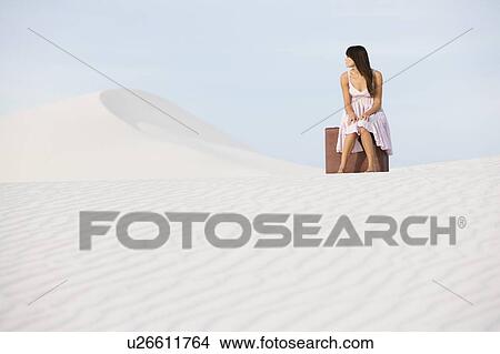 Picture - A young woman sitting on a suitcase in the desert. Fotosearch