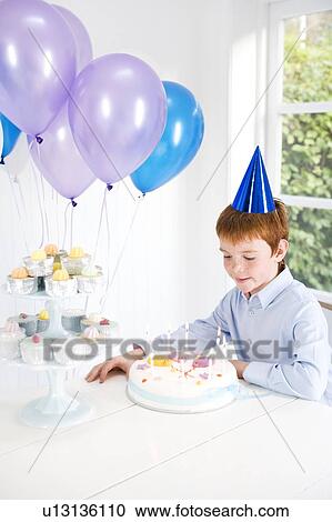 Stock Image - Boy sitting at table with birthday cake and balloons. Fotosearch