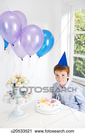 Stock Image - Boy sitting at table with birthday cake and balloons. Fotosearch