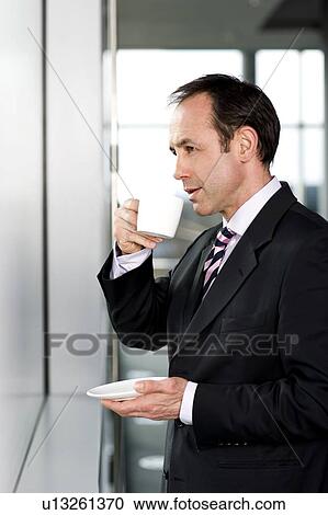 Stock Image - Businessman standing at office window, drinking a cup of coffee. Fotosearch