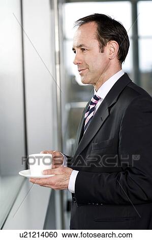 Stock Image - Businessman standing at office window, drinking a cup of coffee. Fotosearch
