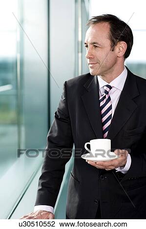 Stock Image - Businessman standing at office window, drinking a cup of coffee. Fotosearch