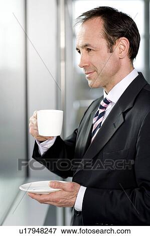 Stock Photo - Businessman standing at office window, drinking a cup of coffee. Fotosearch
