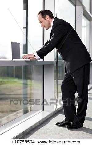 Picture - Businessman standing by a window, typing on a laptop. Fotosearch