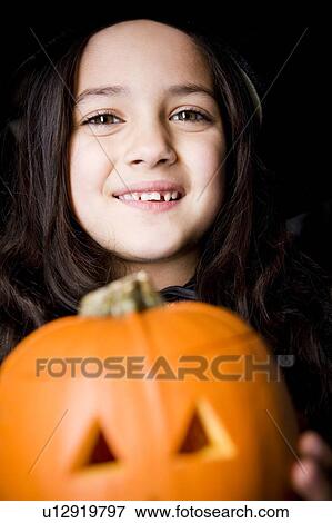 Stock Photo - Girl in costume at a Hallowe'en party, holding a pumpkin with a carved face. Fotosearch