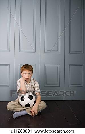 Stock Image - grumpy little boy sitting on floor with football. Fotosearch
