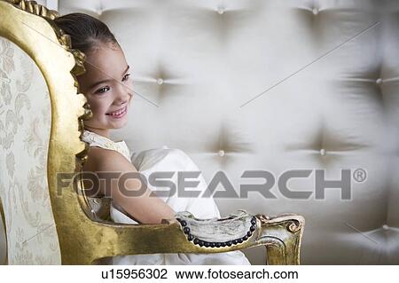 Stock Image - Little girl sitting in an ornate chair wearing a party dress. Fotosearch