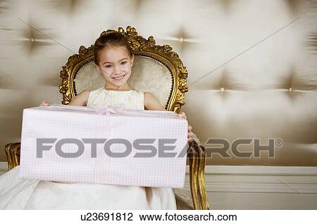Stock Image - Little girl sitting in an ornate chair wearing a party dress holding a large present. Fotosearch
