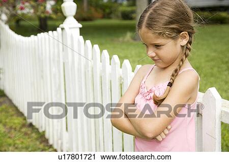 Stock Image - little girl standing against fence with arms folded. Fotosearch