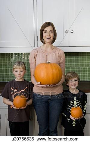 Stock Photograph - Mother and two children in the kitchen holding orange Hallowe'en pumpkins. Fotosearch