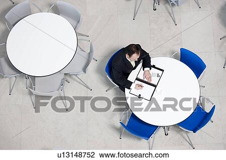 Stock Image - Overhead view of businessman at table of staff canteen, looking through business papers. Fotosearch