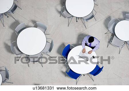 Stock Image - Overhead view of businessman at table of staff canteen, typing on a laptop. Fotosearch