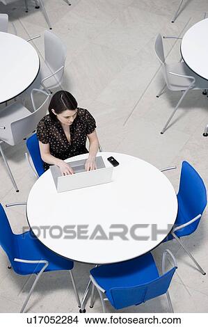 Picture - Overhead view of businesswoman at table of staff canteen, typing on a laptop. Fotosearch