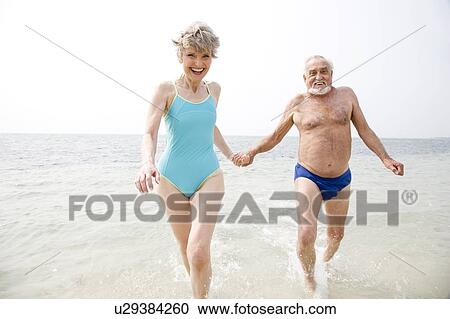 Stock Image - Portrait of a senior couple hand in hand running in the sea. Fotosearch