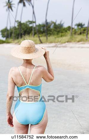 Stock Image - Portrait of a senior woman in a swimsuit at the beach wearing a straw had. Fotosearch