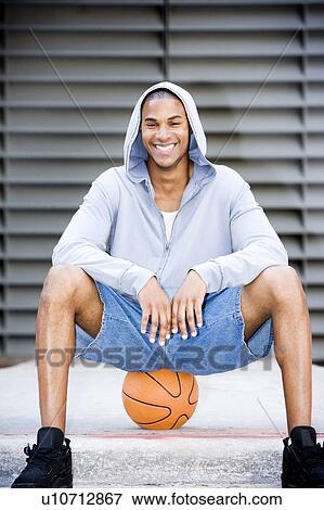 Stock Photo - Portrait of a smiling young African American man in a grey hooded top sitting on a basketball. . Fotosearch
