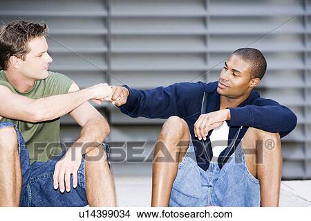 Portrait of a young African American and Caucasian men sitting smiling making greeting hand gestures View Large Photo Image Picture - Portrait of a young African American and Caucasian men sitting smiling making greeting hand gestures . Fotosearch