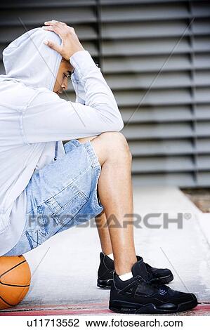 Stock Image - Portrait of a young African American man in a grey hooded top sitting on a basketball, head in hands. . Fotosearch