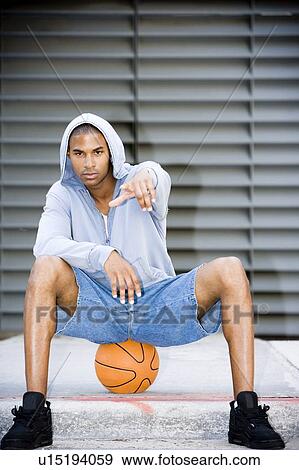 Stock Photo - Portrait of a young African American man in a grey hooded top sitting on a basketball pointing at camera. . Fotosearch