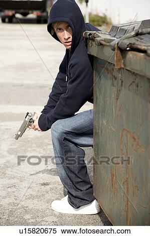 Stock Photography - Portrait of a young man holding a gun sheltering behind a bin . Fotosearch