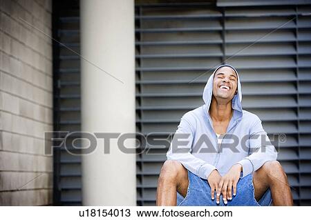 Stock Image - Portrait of a young smiling African American man in a grey hooded top sitting on a basketball. . Fotosearch