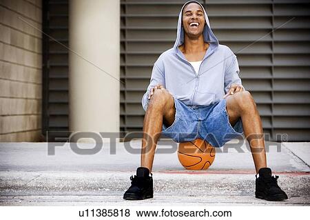 Stock Photo - Portrait of a young smiling African American man in a grey hooded top sitting on a basketball. . Fotosearch