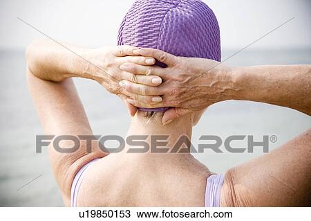 Stock Image - Senior woman in a swimsuit and swimming hat, at the beach looking out to sea. Fotosearch
