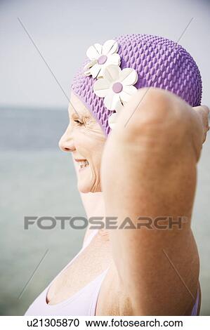 Stock Image - Senior woman in a swimsuit and swimming hat, at the beach looking out to sea. Fotosearch