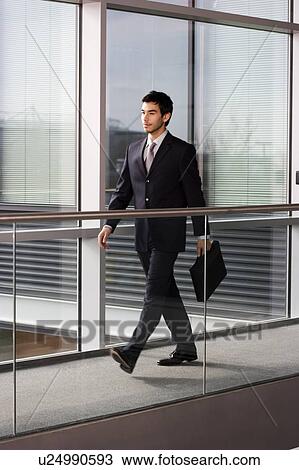 Stock Image - Young businessman in modern office building, walking along with briefcase. Fotosearch