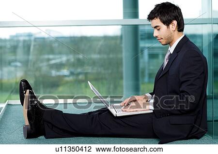 Stock Image - Young businessman sitting on office floor, typing on a laptop. Fotosearch