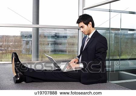 Stock Photo - Young businessman sitting on office floor, typing on a laptop. Fotosearch