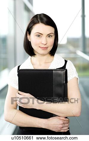 Stock Image - Young businesswoman standing on office walkway holding folder of papers. Fotosearch