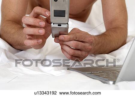 Stock Image - Young man in bed, using mobile phone. Fotosearch