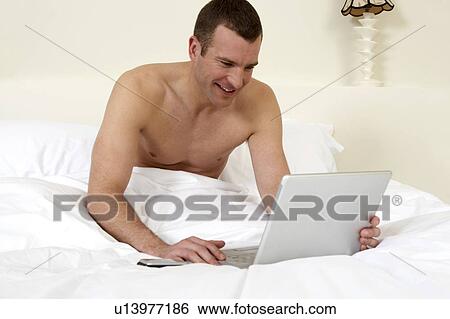Stock Photograph - Young man in bed, working on laptop. Fotosearch