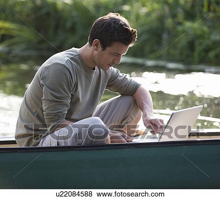 Stock Photo - Young man sitting in a boat working on a laptop. Fotosearch