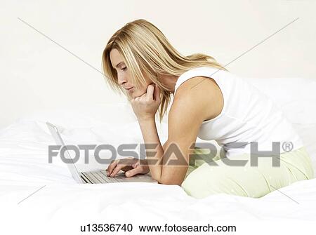 Stock Image - Young woman in bed working on laptop. Fotosearch