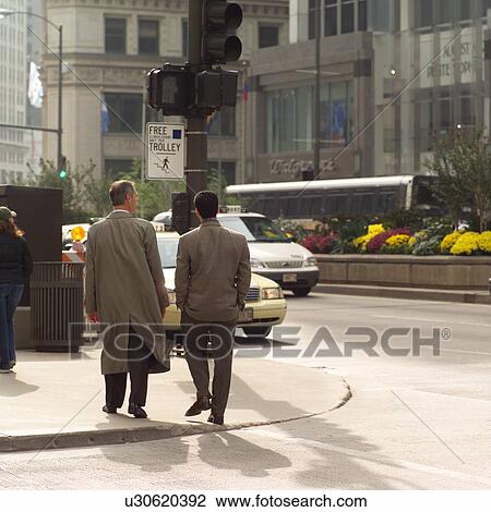 Stock Image - Rear view of two people walking on a sidewalk. Fotosearch