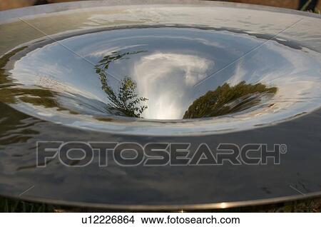 Picture - Metal water fountain in motion with trees and blue sky reflection. Fotosearch