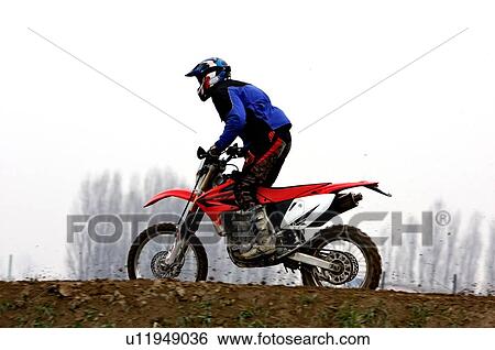 Stock Photograph - Single motocross rider riding along dirt ridge, side view. Fotosearch