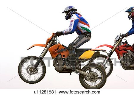 Stock Image - Two motocross riders riding along dirt ridge, side view, front wheels off ground. Fotosearch