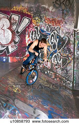 Stock Image - Youths practicing bike wheelies against graffiti wall backdrop. Fotosearch