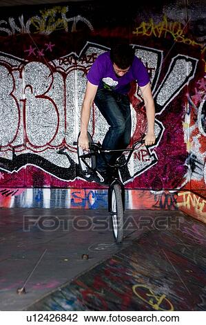 Stock Image - Youths practicing bike wheelies against graffiti wall backdrop. Fotosearch