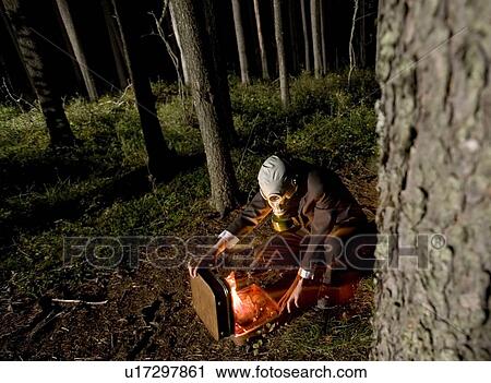 Stock Image - Man in business suit and gas mask opens case with glowing red contents, in eerie forest at night, Cypress Hills. Fotosearch