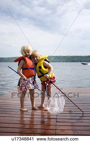 Stock Image - Two young girls wearing life preservers with net and fishing rod embrace, Saskatchewan, Canada. Fotosearch